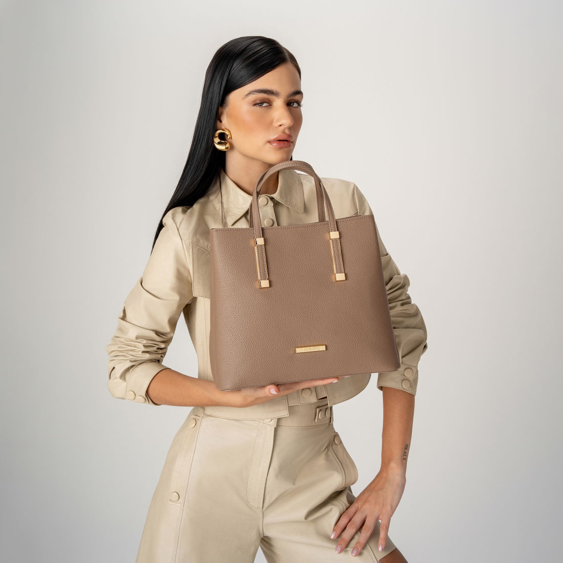 Mulher segurando bolsa tote Via Marte bege com detalhes metálicos dourados
